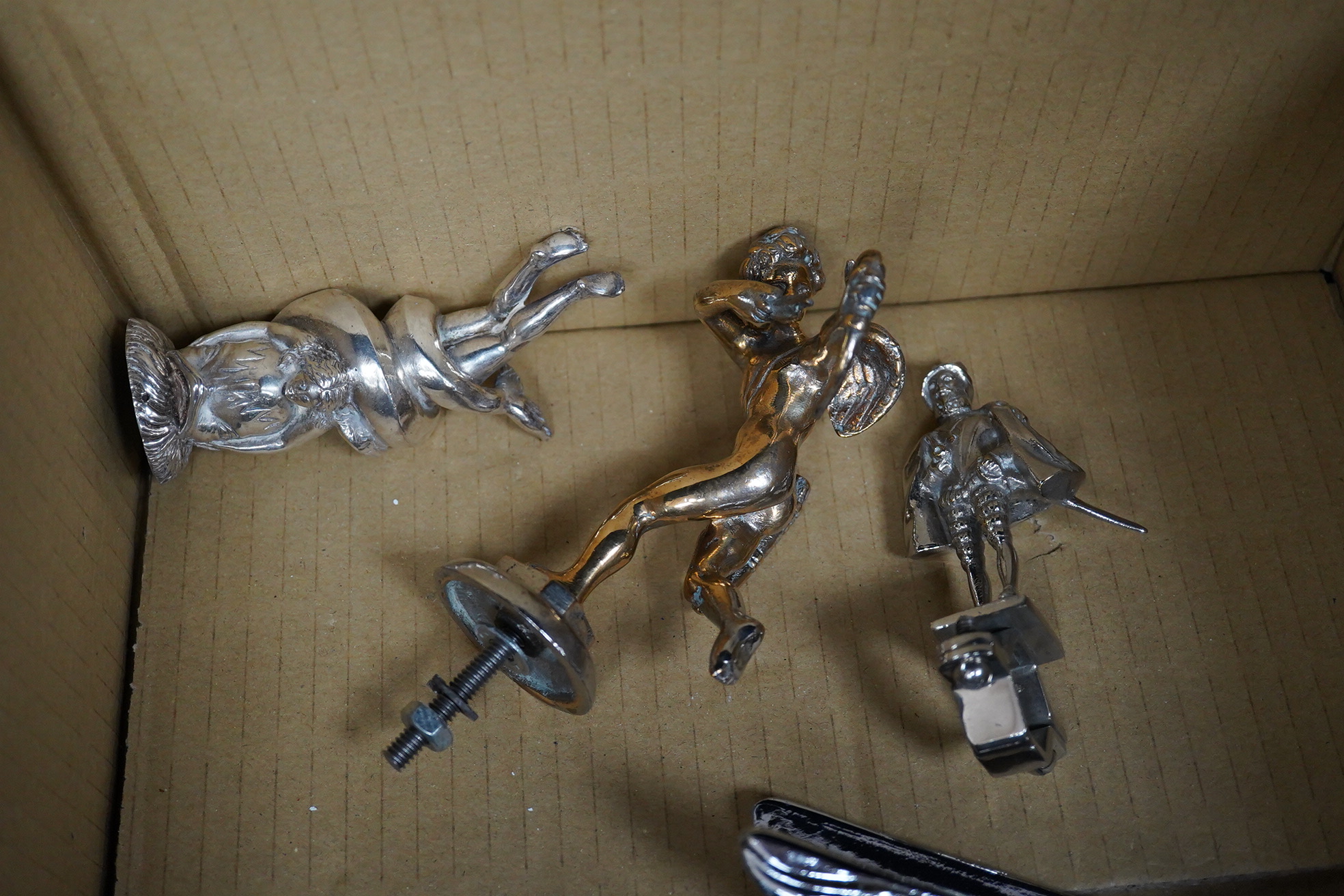 Six chrome and brass car mascots, including; a figure with a dolphin, a figure of Cupid, a grotesque in the form of a devil, a figure of a Tudor gentleman, an Art Deco figure of a flying woman, etc.